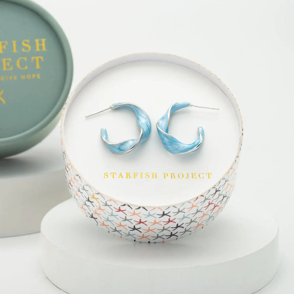 A pair of light blue marbled hoop earrings displayed inside a round white jewelry box with the brand name 'STARFISH PROJECT' printed in gold. The box is set on a white pedestal, with a matching branded round tin partially visible in the background.