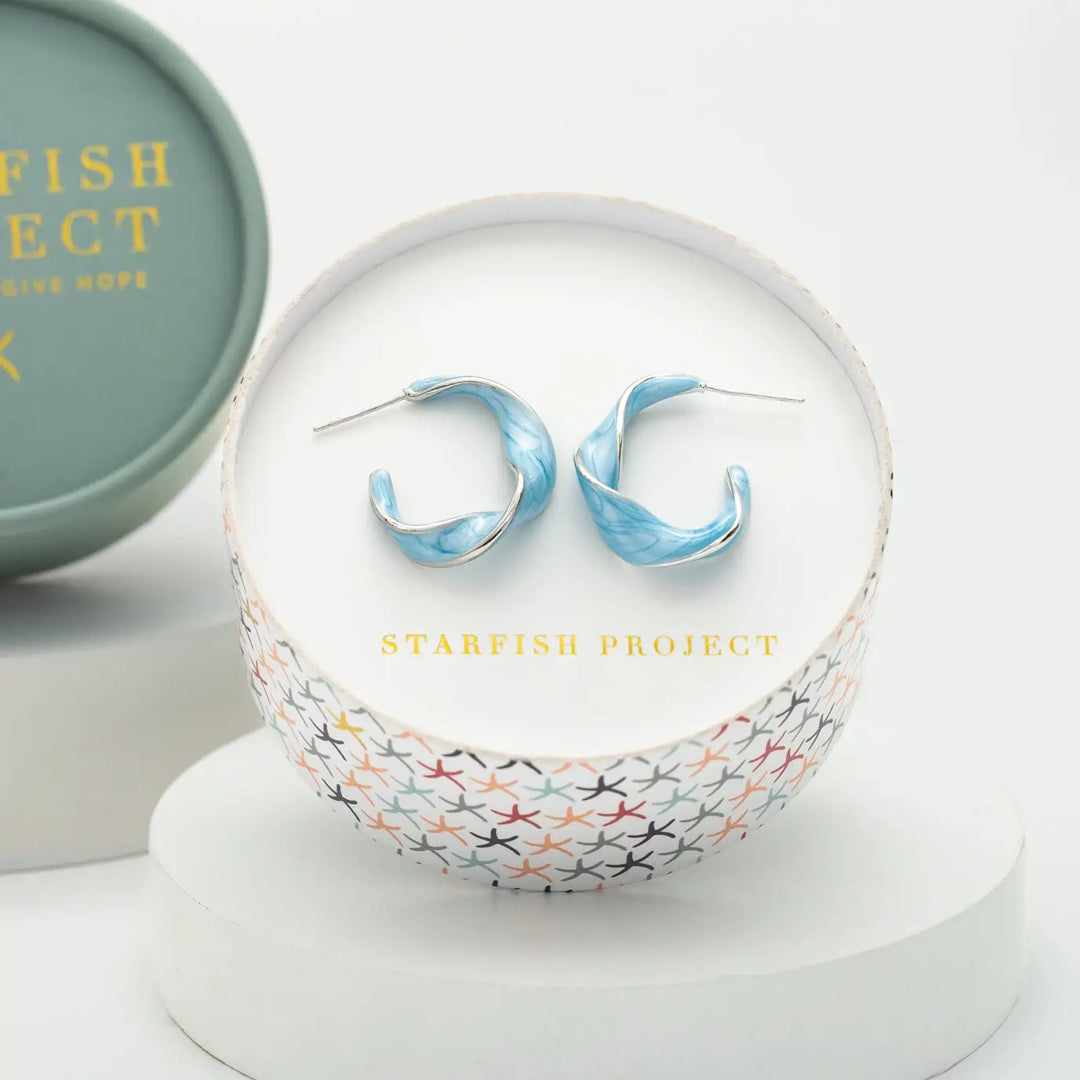 A pair of light blue marbled hoop earrings displayed inside a round white jewelry box with the brand name 'STARFISH PROJECT' printed in gold. The box is set on a white pedestal, with a matching branded round tin partially visible in the background.