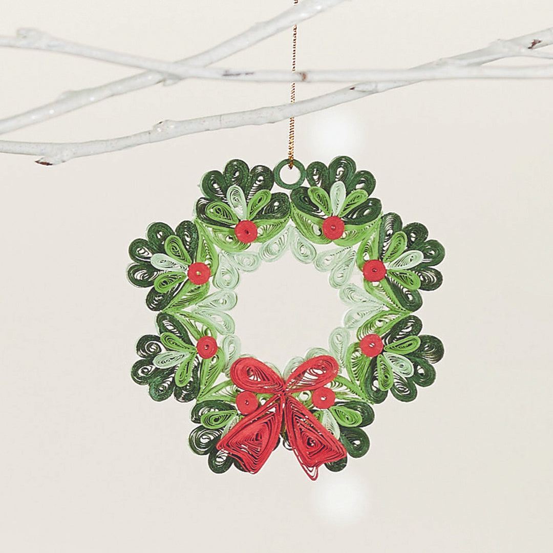 A quilled paper wreath ornament in green with red accents and a red bow, hanging on white branches.