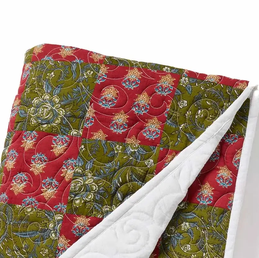 A close-up of a quilted patchwork blanket featuring alternating red and olive green sections with floral and scroll patterns in blue, gold, and beige, with a white quilted reverse side visible.
