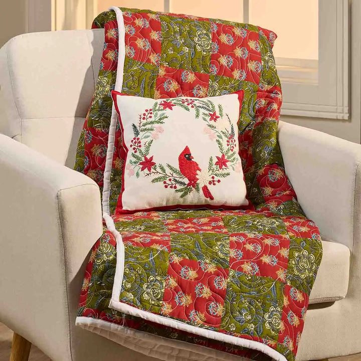 A quilted holiday throw blanket draped over a white armchair, featuring a red and green floral pattern with a white border, accompanied by a matching square pillow with a cardinal design.