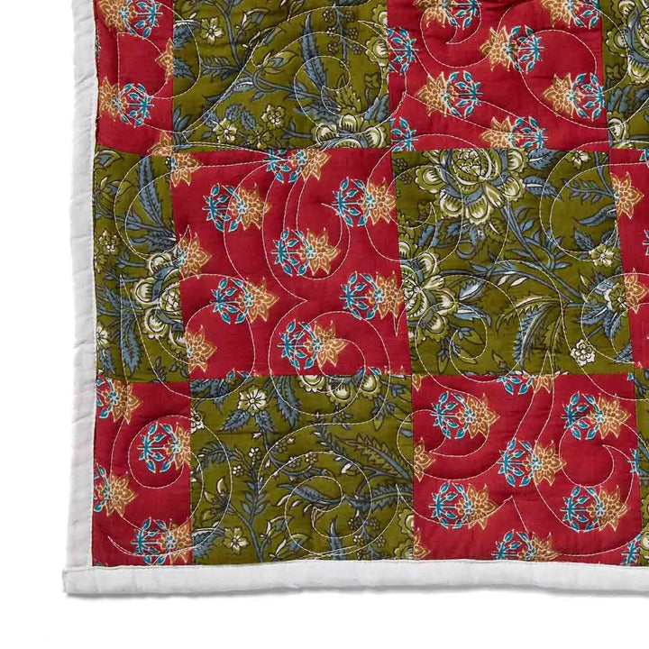 A close-up of a quilted patchwork blanket featuring alternating red and olive green floral panels with blue and gold accents, finished with a white stitched border.