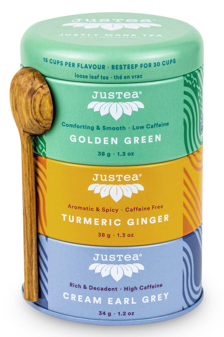 A set of three Justea loose leaf tea tins labeled Golden Green, Turmeric Ginger, and Cream Earl Grey, with a hand-carved teaspoon.