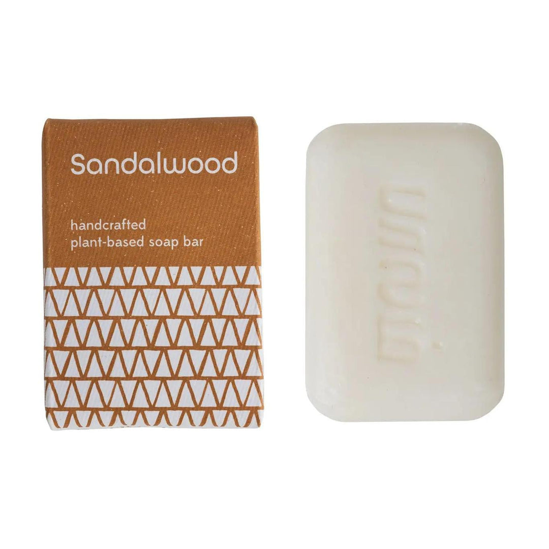 A bar of handcrafted sandalwood soap next to its packaging, which has a geometric pattern with 'SANDALWOOD' and 'Handcrafted in India' text.