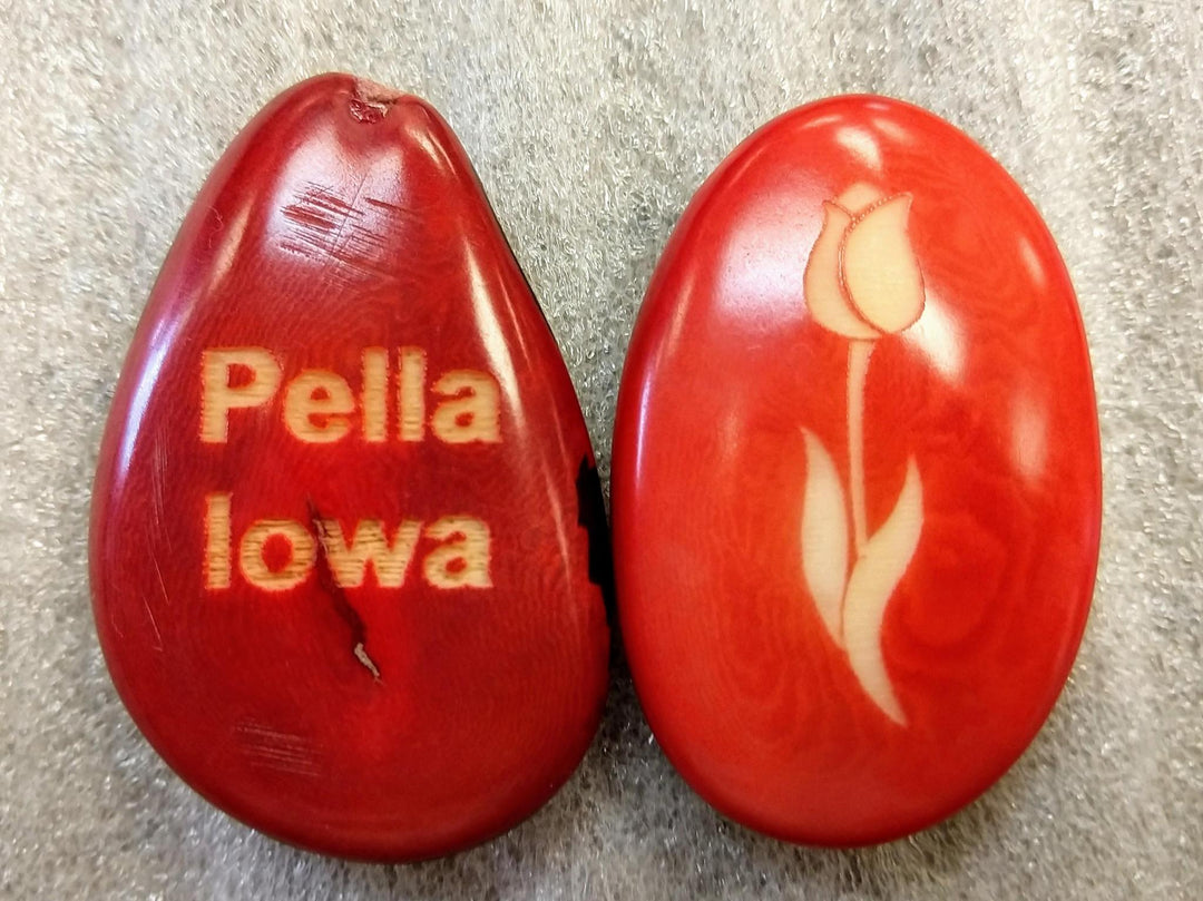 Two polished red tagua nuts with the text 'PELLA, IOWA' carved on them, displayed against a grey background.