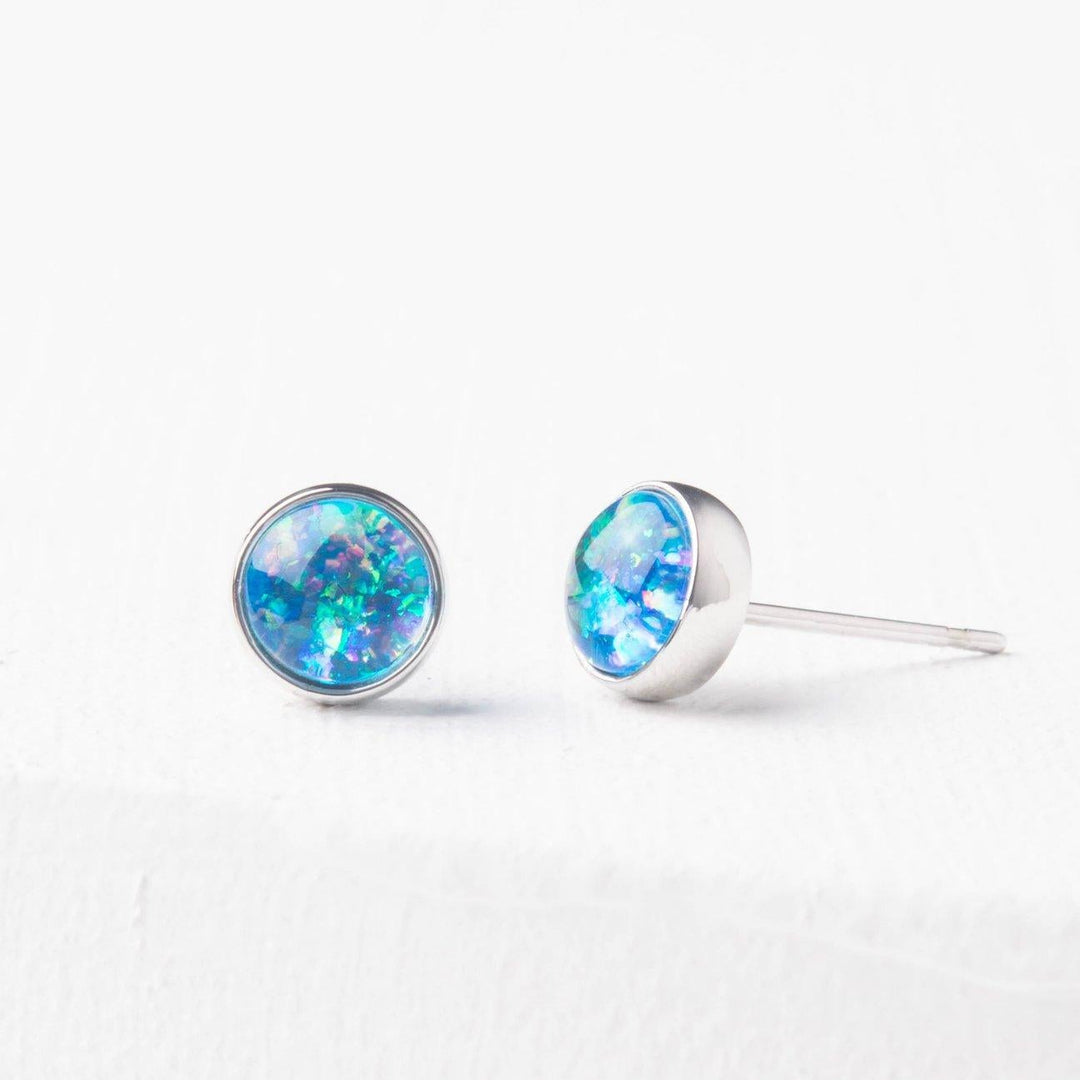 A pair of blue and silver stud earrings with a sparkling opal centerpiece, placed on a white surface.