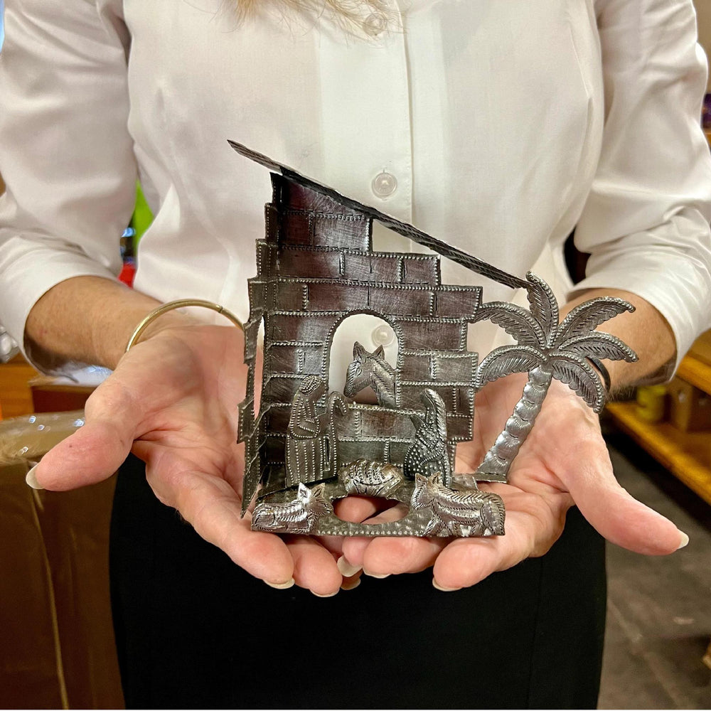 Lady holding nativity scene under a brick house with a palm tree and animals surrounding Joseph, Mary, and Jesus. 