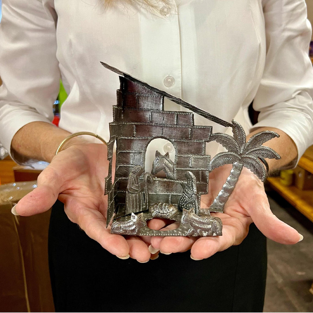 Lady holding nativity scene under a brick house with a palm tree and animals surrounding Joseph, Mary, and Jesus. 