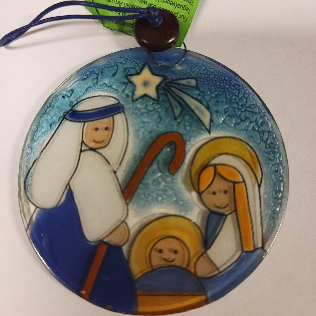 A round glass nativity scene ornament with a depiction of the three smiling faces of Mary, Joseph, and baby Jesus surrounded by a starry night background. The ornament has a blue hue and is adorned with recycled glass and water-based paint.
