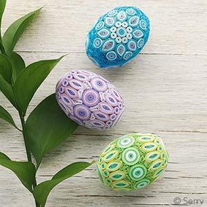 Three quilled paper Easter eggs in blue, purple, and green, displayed on a wooden surface with a green leafy branch to the side.