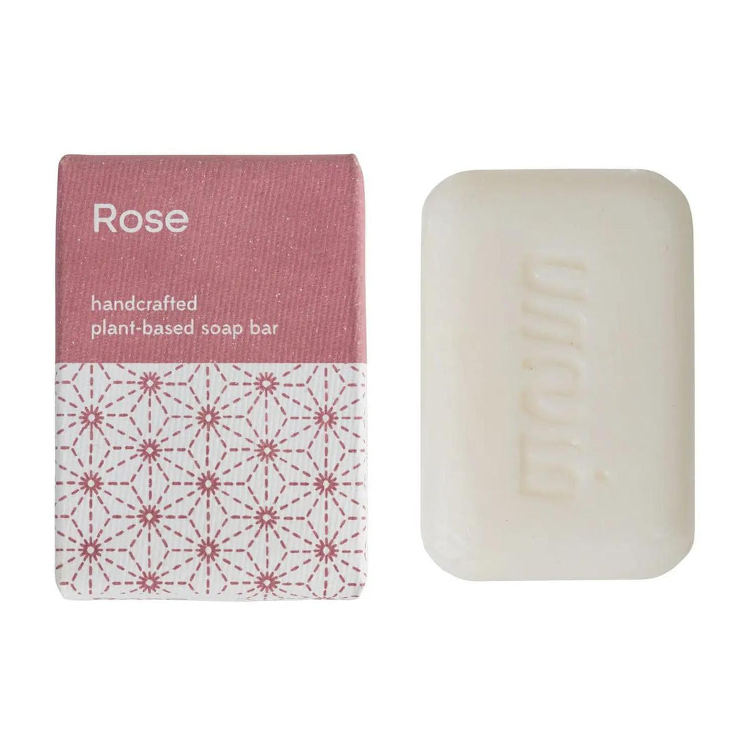 A bar of soap with a pink label that reads 'rose' and 'handcrafted bar soap made with love in India'. The packaging is predominantly pink and white with a floral pattern.