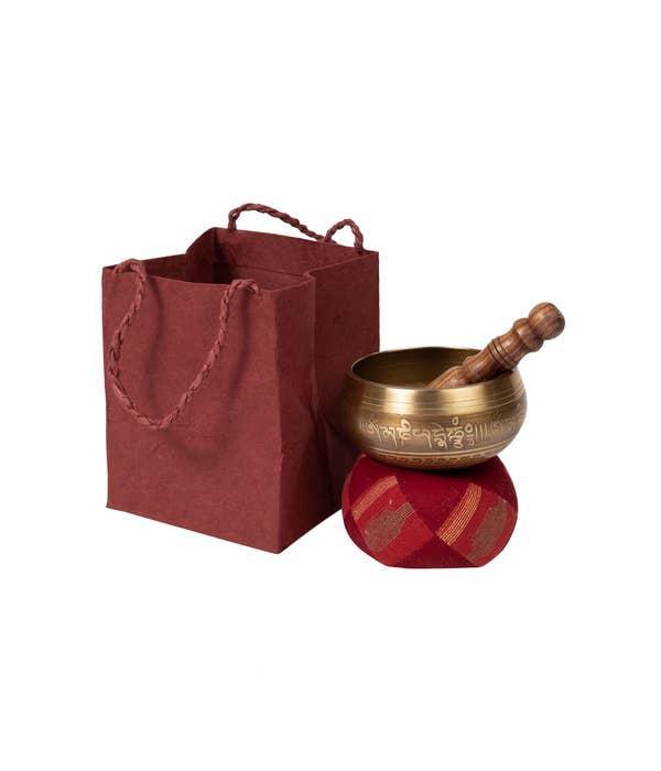 A handcrafted brass singing bowl with a rosewood striker and a red gift bag with a twisted cord handle.