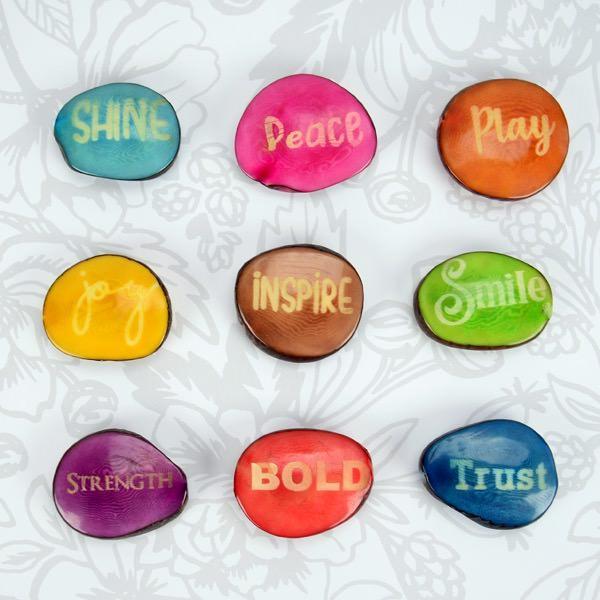 A collection of polished tagua nuts each dyed a different color and inscribed with a word, such as 'Peace,' 'Play,' and ' Inspire.'.