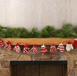 A garland of red and white wool mittens hanging over a fireplace, with a mantle above it decorated with green pine branches.